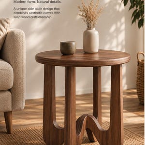 May include: A round wooden side table with a dark brown finish, featuring three curved legs that connect at the base. On the table surface sits a small dark gray bowl and a light-colored ceramic vase containing dried wheat-like stalks.