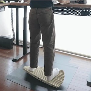 May include: A light-coloured wooden balance board with a ribbed, fan-shaped surface. The board features a series of small, dark circular fasteners along its side edge, designed for standing on while working at a desk to encourage movement.