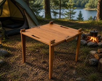 May include: A square, wooden camping table with a light brown finish. The table has four legs and a folding design. It is set up outdoors near a tent, campfire, and lake.