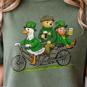 May include: Green t-shirt with a St. Patrick's Day graphic. The graphic features a duck, a bear, and a leprechaun riding a tandem bicycle. The duck is wearing a green suit and hat, the bear is wearing a green sweater and cap, and the leprechaun is holding a beer.