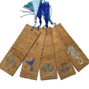 May include: Set of five cork bookmarks with scalloped edges and embroidered sea-themed designs. Each bookmark features a different design, including a seashell, seahorse, and whale tail. The bookmarks are topped with blue and teal tassels.
