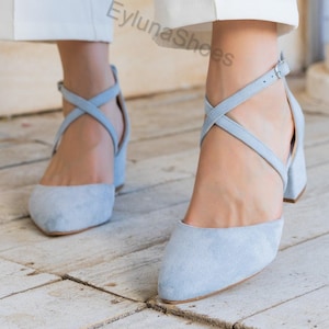 以下が含まれることがあります： ライトブルーのスエードヒール。足首にクロスストラップ、ブロックヒール。靴はポインテッドトゥでバックル留め。画像には「EylunaShoes」の文字が見えます。
