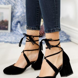 May include: Black suede heels with a block heel and ankle ties. The shoes have a pointed toe and a strap that wraps around the ankle. The heels are approximately 2 inches high.