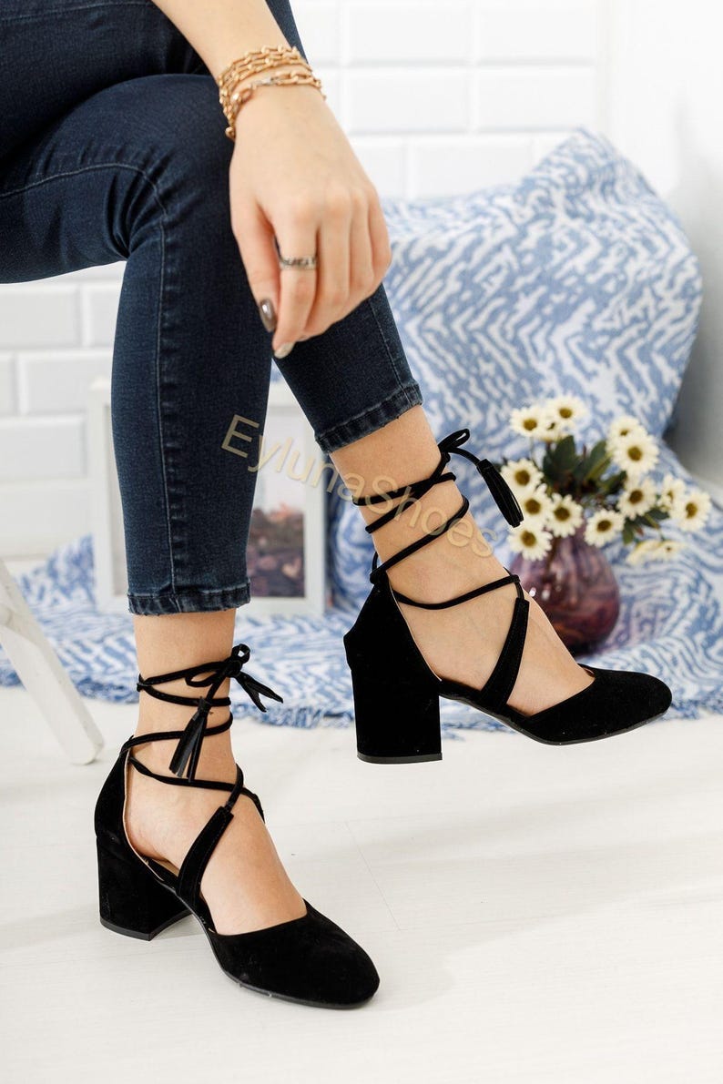 May include: Black suede ankle strap heels with a block heel. The shoes feature a crisscross strap design and tie closure with tassels. The heels are approximately 3 inches high.