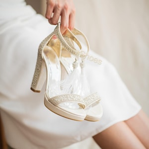 May include: Ivory-colored platform heels with rhinestone embellishments. The heels have a thick block heel and a strap around the ankle. White feathers dangle from the ankle strap. The shoes are held up by a person.