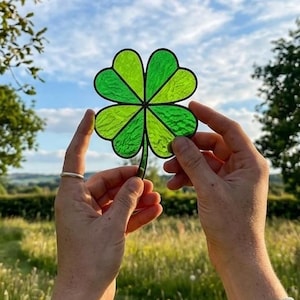 May include: A four-leaf clover suncatcher crafted from translucent green stained glass with dark soldered metal seams. The clover features four distinct heart-shaped leaves and a short stem, allowing light to pass through the textured glass surface.