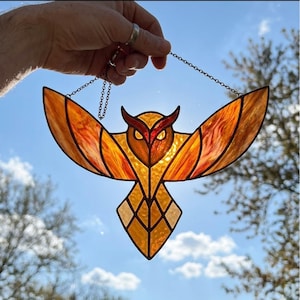 Peut inclure: Suncatcher en vitrail en forme de hibou aux ailes déployées. Le hibou présente des nuances d'orange, de jaune et de rouge, avec une chaîne pour l'accrocher. Le design est tenu par une main contre un ciel bleu nuageux.