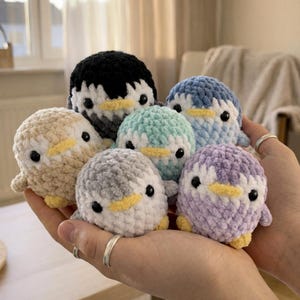 May include: A collection of handmade crochet penguin plushies in various colors, including black, blue, gray, beige, teal, and purple. Each penguin has a yellow beak and black eyes. The plushies are soft and cuddly, perfect for a child's toy collection.