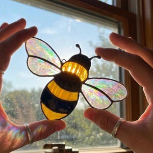 May include: A stained glass bee suncatcher featuring iridescent wings that shimmer with rainbow colors. The body of the bee is constructed from alternating segments of opaque yellow and deep black glass, with a dark glass head and two antennae.