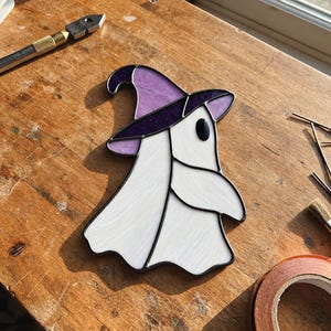 May include: A stained glass ghost ornament featuring a white body with a subtle texture, wearing a purple witch hat with a dark purple band. The piece is constructed with black solder lines and includes a single black oval eye detail.