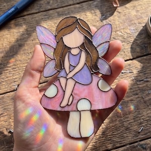 May include: A stained glass suncatcher featuring a figure with brown hair and a purple dress sitting on a pink mushroom with white spots. The figure has iridescent wings and is constructed with dark metal solder lines between the glass pieces.