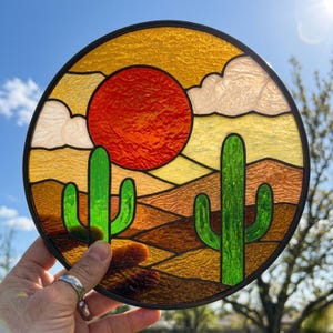 Op de afbeelding: Rond glas-in-lood kunstwerk met een woestijnlandschap. Het ontwerp omvat een felrode zon, groene cactussen en een gelaagde achtergrond in tinten geel, oranje en bruin. Het kunstwerk is omlijst met een zwarte rand.