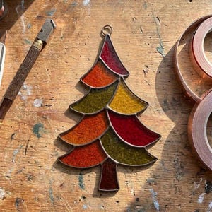 May include: A stained glass Christmas tree ornament featuring a multi-colored mosaic design with segments in shades of red, orange, yellow, and olive green. The piece is constructed with dark metal solder lines and includes a small circular metal loop at the top.