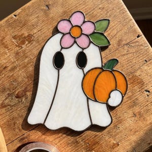 Op de afbeelding: Een glas-in-lood spookornament met een wit lichaam, twee zwarte ovale ogen en een roze bloem met een oranje hart op het hoofd. Het spook houdt een feloranje pompoen vast met een groene steel en een klein wit accent aan de zijkant.