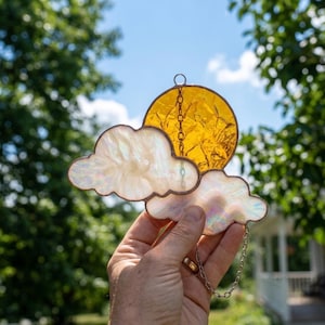 Puede incluir: Atrapasoles de vidriera con un sol amarillo y dos nubes blancas iridiscentes. El sol es de vidrio ámbar texturizado y las nubes tienen un brillo nacarado. Una cadena plateada está unida para colgarlo. Una pieza decorativa.