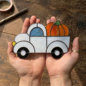 May include: A stained glass ornament shaped like a white pickup truck with a blue window, a yellow headlight, black wheels, and a large orange pumpkin in the truck bed. The piece is constructed with dark metal solder lines between the glass segments.