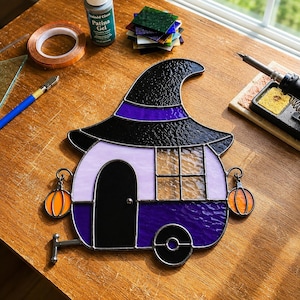 May include: A stained glass suncatcher shaped like a camper trailer topped with a black witch hat. The camper features light purple and dark purple glass panels, a black door, a grid window, and two small orange pumpkin accents on the sides.