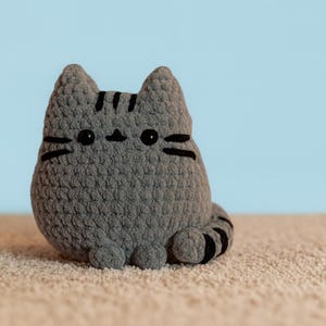 Puede incluir: Un peluche de gato gris hecho a mano con ganchillo, con rayas negras y detalles faciales. El gato tiene un cuerpo redondeado, orejas pequeñas y una cara sonriente. El peluche está sobre una superficie beige con un fondo azul claro.