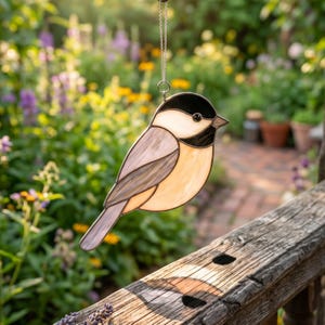 Könnte beinhalten: Ein Sonnenfänger aus Buntglas in Form einer Meise mit schwarzem Kopf, weißem Wangenfleck und hellbraunem Körper mit grauen Flügeldetails. Der Vogel ist an einer dünnen silbernen Metallkette aufgehängt und hat eine glatte Glasoberfläche.