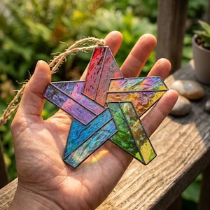 May include: A five-pointed star ornament crafted from iridescent stained glass pieces in shades of red, orange, yellow, green, blue, and purple. The glass segments are joined by dark metal solder lines and include a natural jute twine hanging loop.