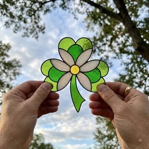 May include: A stained glass suncatcher shaped like a three-leaf clover with a central white flower. The clover features vibrant green glass segments, while the flower has white petals and a small yellow circular centre, all joined by dark metal lines.
