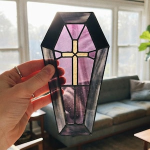 May include: A coffin-shaped stained glass piece featuring translucent purple glass panels framed in black metal. A central cross made of textured, light-colored glass is integrated into the design, creating a geometric and religious motif.