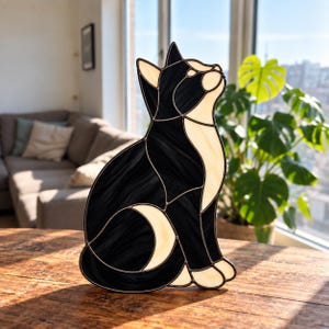May include: A stained glass cat figure constructed from black and cream-coloured glass pieces joined by dark metal solder lines. The cat is depicted in a seated position, looking upward with its head tilted, featuring a two-tone colour pattern.