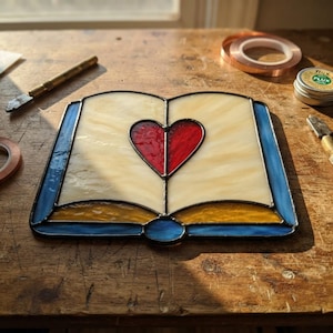 Op de afbeelding: Een glas-in-lood suncatcher in de vorm van een open boek met een rood hart in het midden. De pagina's zijn gemaakt van crèmekleurig glas, de onderranden zijn amberkleurig en de buitenrand is blauw. De delen zijn verbonden door donkere metalen soldeerlijnen.