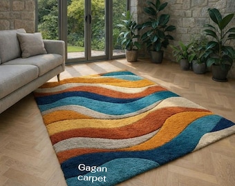 May include: A rectangular area rug with a wavy pattern in shades of blue, orange, yellow, and beige. The rug is placed on a wooden floor in a room with a gray sofa and large windows. The text "Gagan carpet" is visible.