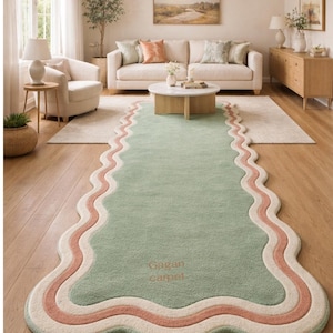Puede incluir: Una alfombra verde con bordes ondulados y un borde rosa y blanco, colocada en un salón. La alfombra, con el texto "Gagan Carpet", añade un acento suave y colorido al suelo de madera. La habitación cuenta con un sofá blanco, una mesa de centro redonda y una lámpara colgante tejida.