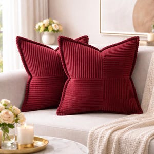May include: Two burgundy corduroy throw pillows with a decorative stitched edge rest on a cream-colored sofa. One pillow has a patchwork design. The pillows add a touch of texture and color to the living room.