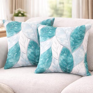 May include: Two square throw pillows with a leaf pattern. The leaves are in shades of teal, light blue, and white, with silver-gray outlines. The pillows are on a white sofa, creating a fresh, nature-inspired aesthetic.