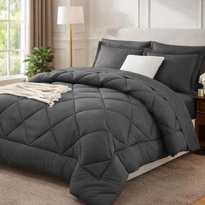 May include: A bed is made with a dark gray comforter and matching pillowcases. A white decorative pillow and a cream-colored throw blanket are also on the bed. The bed is in a well-lit room with a neutral color scheme.