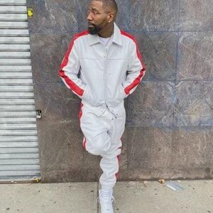 May include: A light gray tracksuit with red stripes on the sleeves and sides. The jacket has a collared neckline and pockets. The matching pants are cuffed at the ankles. The person is wearing white sneakers.