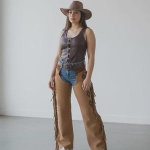 May include: A brown leather vest with stud details and a matching cowboy hat. The outfit includes denim jeans, brown suede chaps with fringe, and a brown belt. White sneakers complete the western-inspired ensemble.