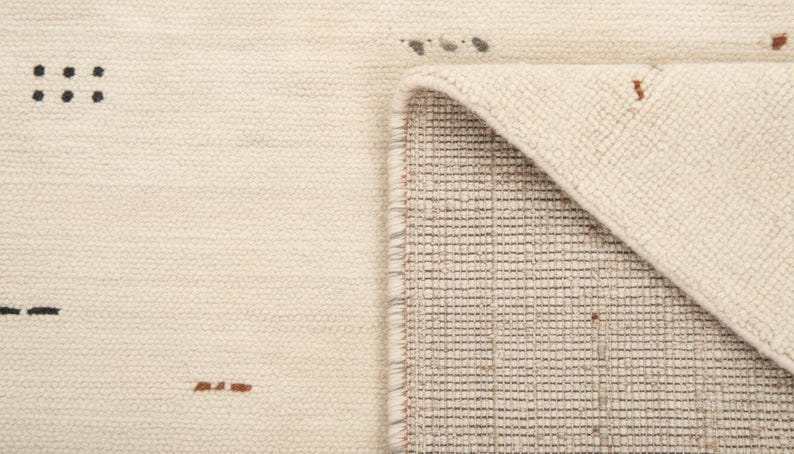 May include: Cream-colored woven rug with a geometric design. The rug features small black and brown accents, with a folded corner revealing the underside's grid pattern. The texture appears soft and inviting, suitable for home decor.