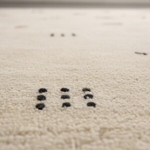 May include: Close-up of a cream-colored rug with a textured surface. The rug features a pattern of small, dark dots and dashes scattered across the surface. The dots are arranged in small groups.