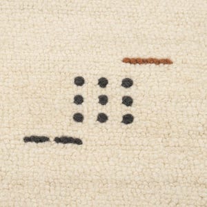 May include: Close-up of a cream-colored rug with a textured surface. The rug features a pattern of dark gray dots arranged in a grid, along with a brown horizontal line and a shorter dark gray line.