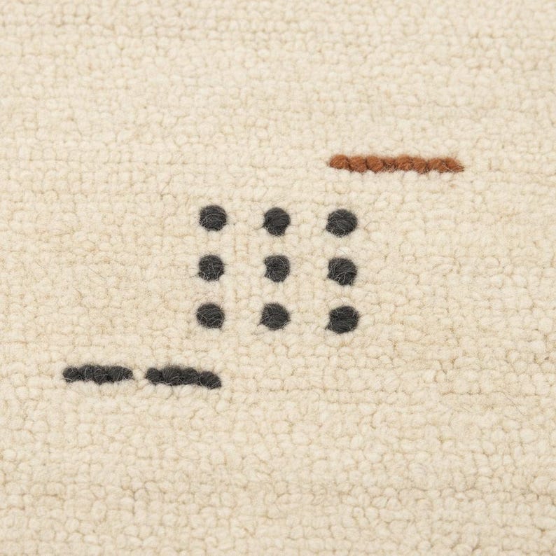 May include: Close-up of a cream-colored rug with a textured surface. The rug features a pattern of dark gray dots arranged in a grid, along with a brown horizontal line and a shorter dark gray line.