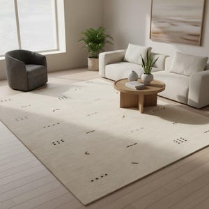 May include: A cream-colored rectangular area rug with a minimalist design. The rug features scattered dark brown geometric shapes, including dots, dashes, and angled lines. The rug is placed in a living room setting with a sofa, chair, and coffee table.