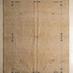 May include: A rectangular area rug in a solid beige color featuring a thin, dark-colored border composed of dotted lines. The design includes small geometric symbols such as diamonds, chevrons, and crosses placed along the perimeter and center.