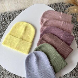 Könnte beinhalten: Eine Auswahl an Strickmützen in Pastellfarben, darunter Gelb, Lavendel, Grün und Rosa. Die Mützen sind auf einer weißen Oberfläche mit umgeschlagenem Saum und Rippstruktur ausgestellt. Die Mützen sind im lässigen Stil gehalten.