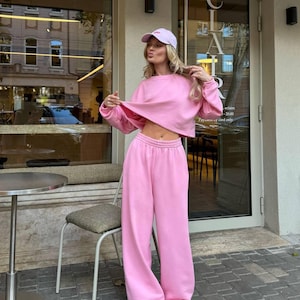 Pode incluir: Um conjunto de fato de treino rosa claro de duas peças. O conjunto inclui um moletom curto com gola redonda e calças de fato de treino de perna larga. A modelo também usa um boné de beisebol rosa claro e tênis brancos. O fundo inclui uma porta de vidro com a palavra "CLAIR" visível.