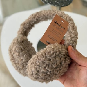 May include: A pair of beige earmuffs with a fluffy, textured appearance. A small tag is attached, with the words "Thank you for your order" printed on it. The earmuffs are designed to keep ears warm in cold weather.