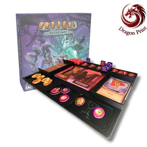 Puede incluir: Un juego de mesa titulado Brzdęk! Katakumby con una caja morada que presenta una ilustración de un dragón. El juego incluye una bandeja organizadora de plástico negro con varias fichas circulares, cartas rectangulares y marcadores rojos.