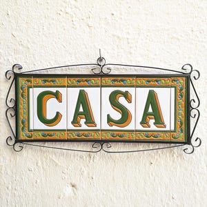May include: Rectangular sign with the word "CASA" in green and orange letters on white tiles. The tiles are framed with a decorative border and set in a black wrought iron frame. The sign hangs against a textured white wall.