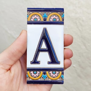 Op de afbeelding: Rechthoekige keramische tegel met de letter "A" in donkerblauw op een witte achtergrond. De tegel heeft een decoratieve rand in blauw, geel en roze. De tegel wordt in een hand gehouden.