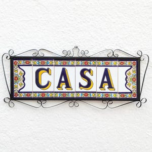 May include: A rectangular sign with the word "CASA" in yellow and blue letters. The sign is made of white tiles with floral and geometric patterns. It is framed in black metal with decorative scrollwork.