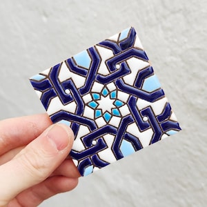 May include: Square ceramic tile with a geometric pattern in white, navy blue, and light blue. The design features interlocking lines and a central starburst motif. The tile is held by a hand.