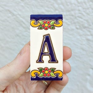 Pode incluir: Uma telha de cerâmica retangular com a letra "A" em azul marinho. A letra está centrada em um fundo branco. As bordas superior e inferior apresentam uma borda azul marinho com desenhos florais amarelos, vermelhos e verdes. A telha é segurada por uma mão.